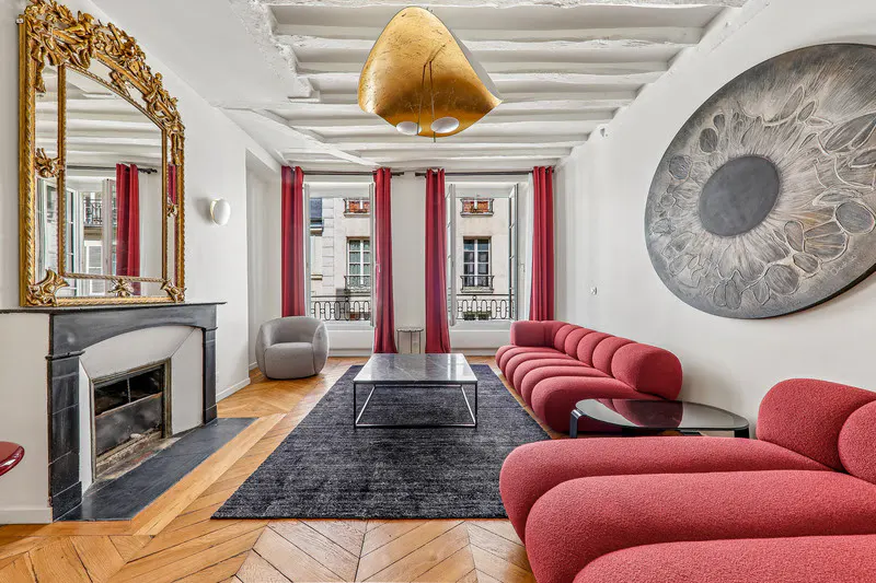 Living room with red sofa, black rug, and fireplace with gold mirror. Windows with red curtains show city view.