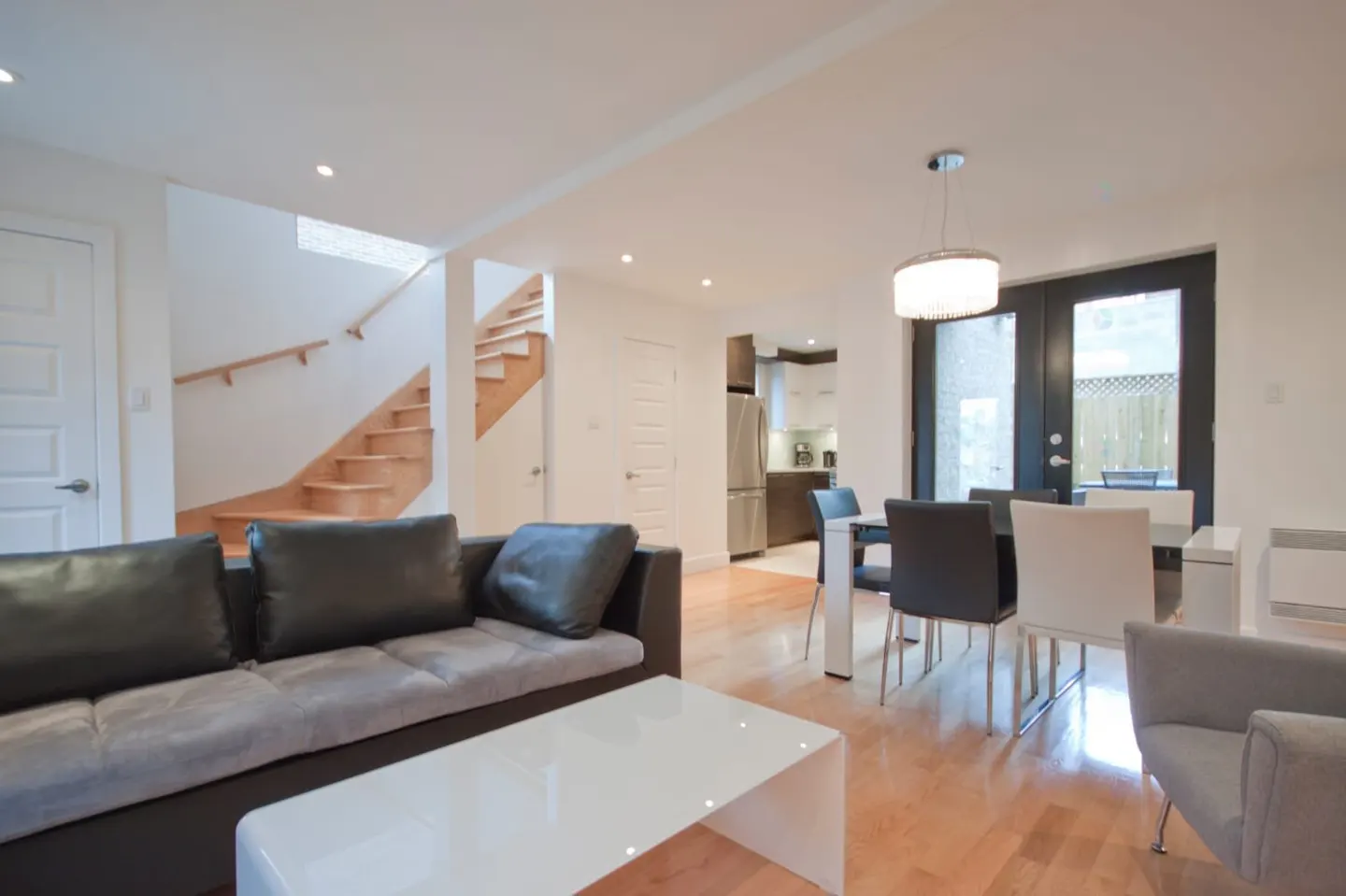 Modern open-concept living area with a black sofa, white coffee table, dining set, and wooden staircase in the background.