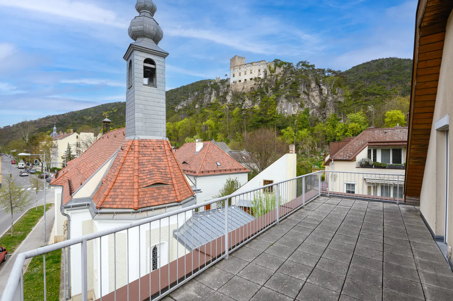 View from a balcony with gray tiles and railings, overlooking a church with a red tile roof and a castle on a hill.