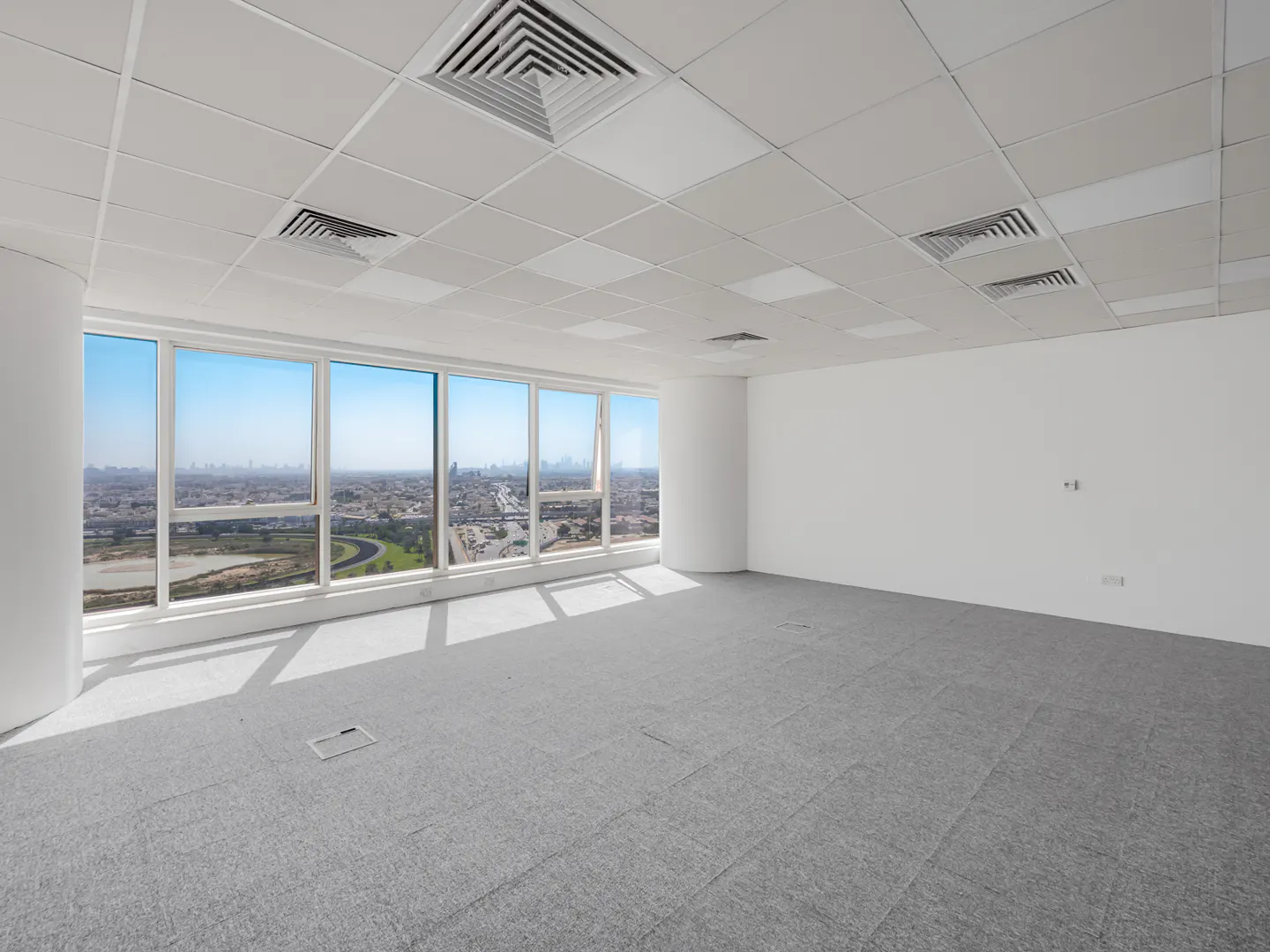 Bright, empty office space with gray carpet, white walls, and large windows overlooking a city skyline under a blue sky.