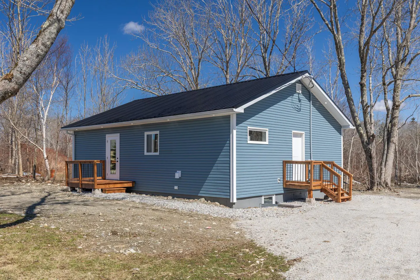 Exterior view of a blue house with a black roof, two wooden decks, and leafless trees against a blue sky.