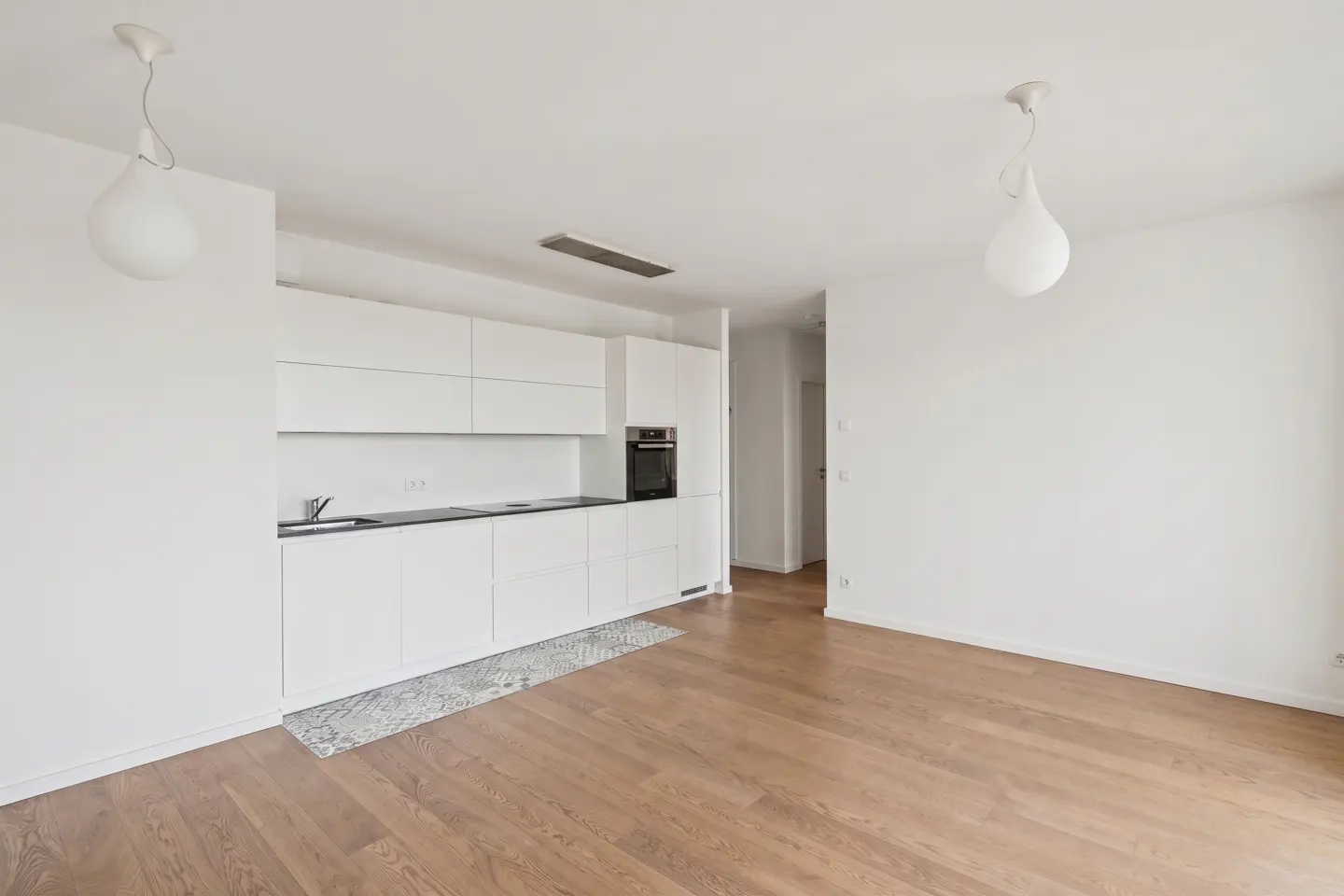 Bright, open-concept apartment with white walls, wood floors, and a modern white kitchen with black countertops. Two pendant lights hang from the ceiling.