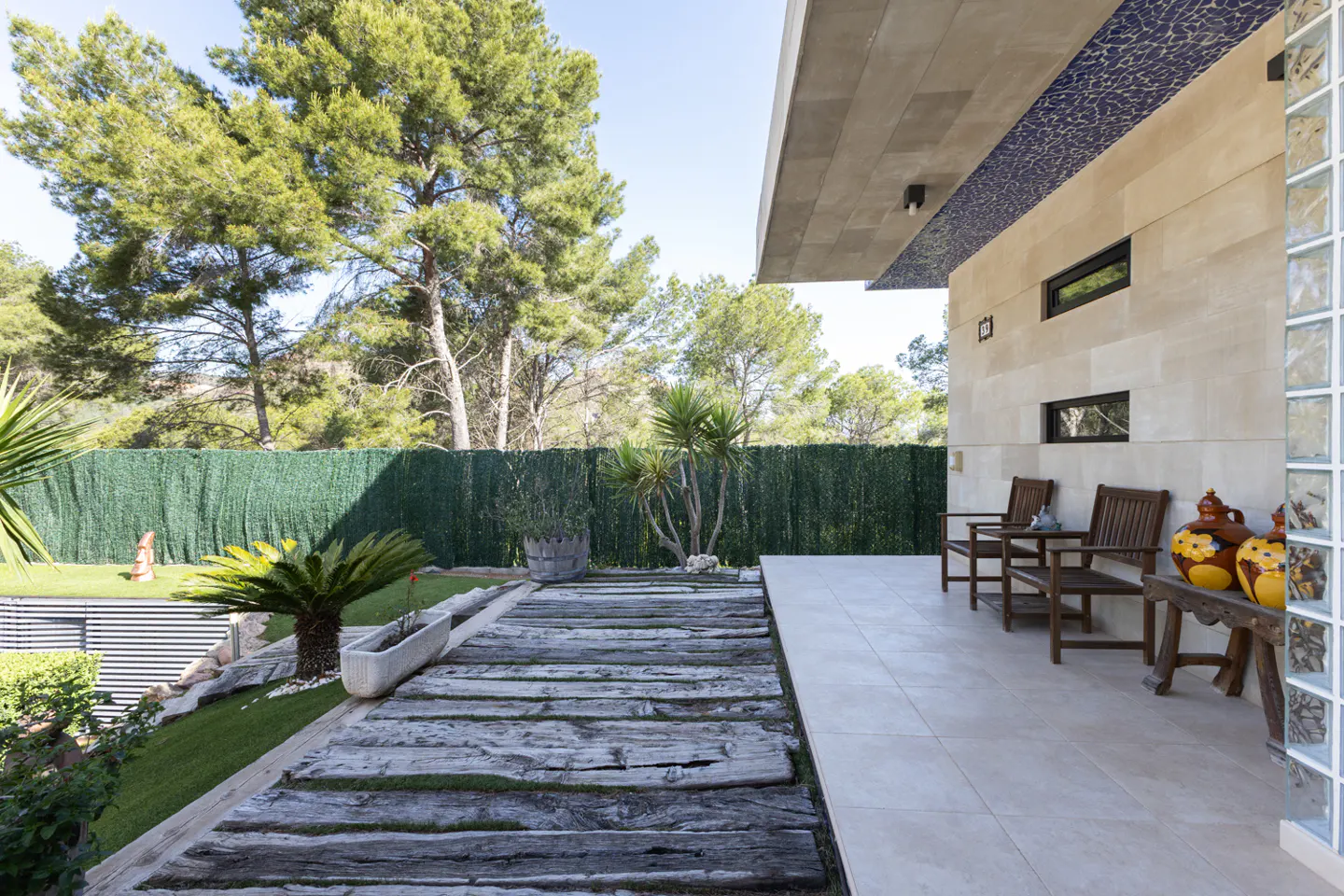 Exterior view of a modern home with a stone patio, wooden benches, and a garden with trees and a green hedge.