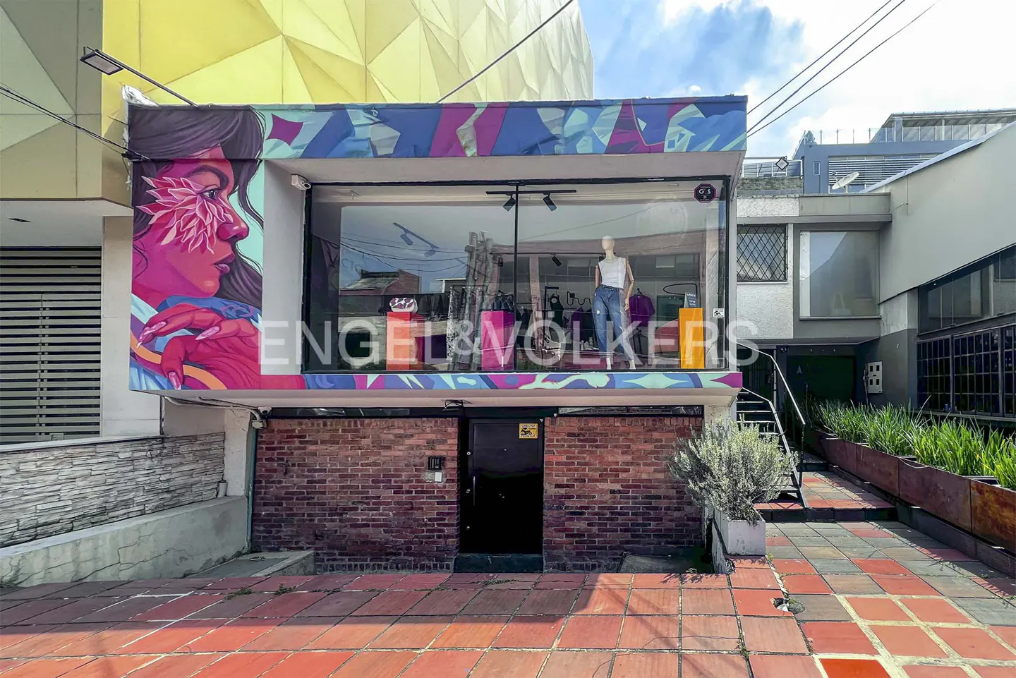 Engel & Volkers real estate office with colorful mural and mannequin in window. Red brick facade and tiled walkway.