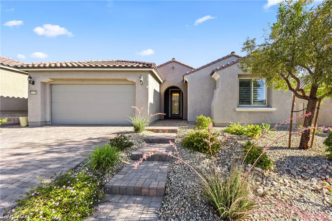 Beige single-story house with a brown tile roof, a gray garage door, and a stone pathway leading to the front door. Landscaped yard with rocks and plants.