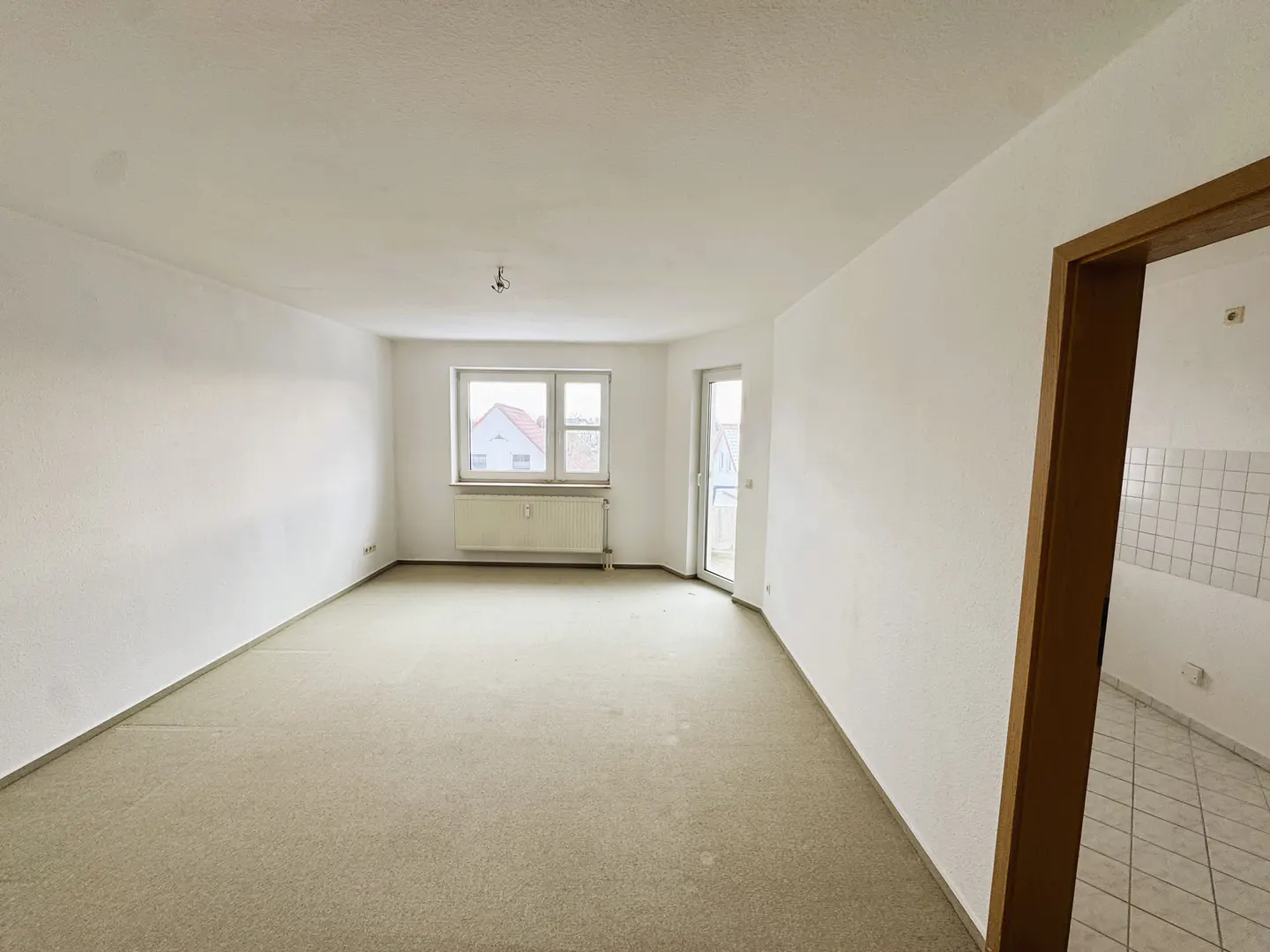 Empty room with beige carpet, white walls, window, radiator, and door to a balcony. A doorway leads to a tiled room.