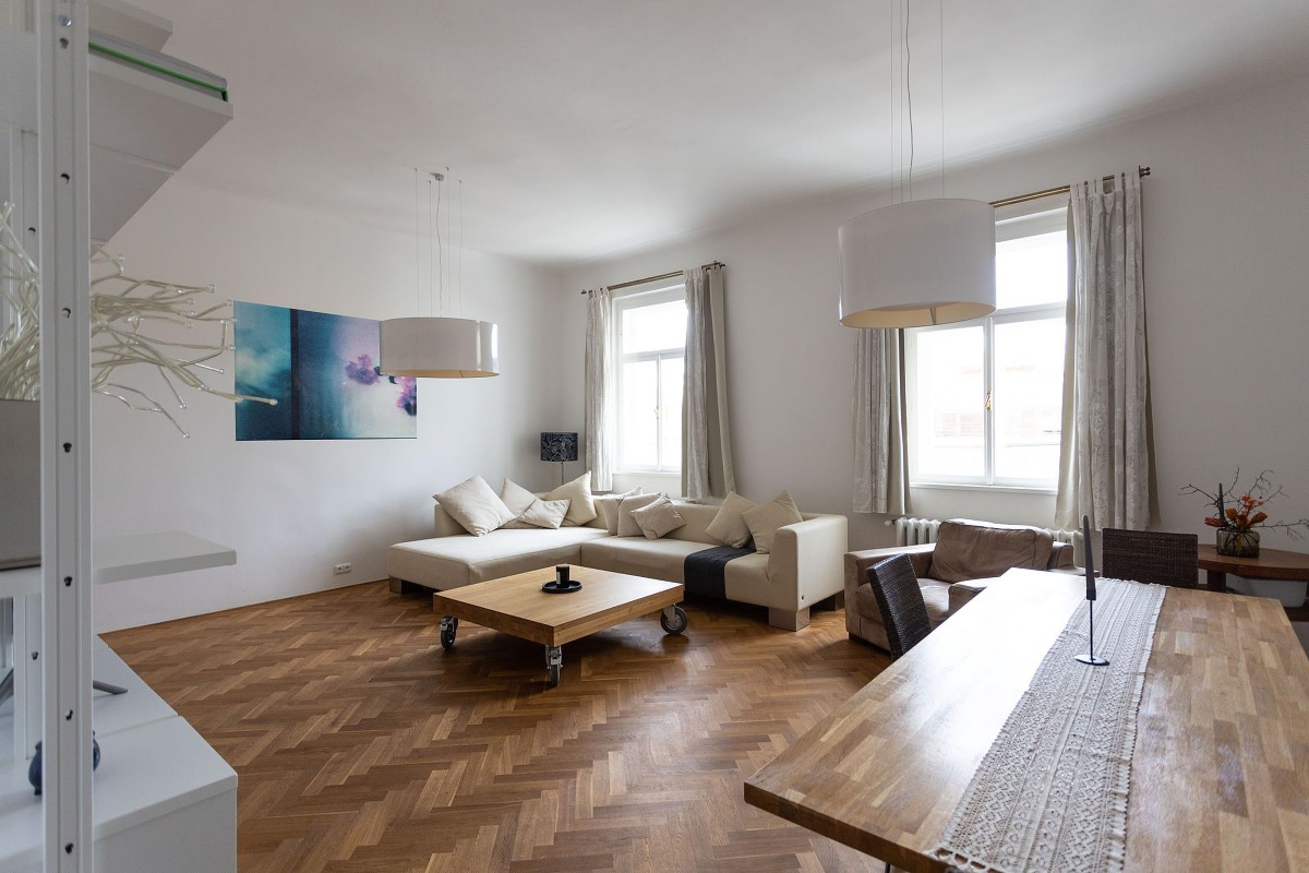 Bright living room with herringbone wood floors, a beige sectional sofa, and a wooden dining table. White walls and pendant lights.