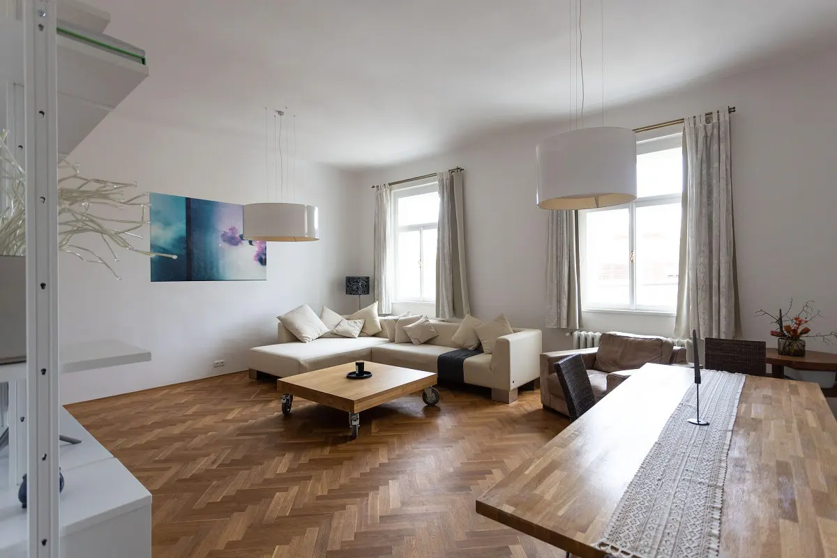 Bright living room with herringbone wood floors, a beige sectional sofa, and a wooden dining table. White walls and pendant lights.