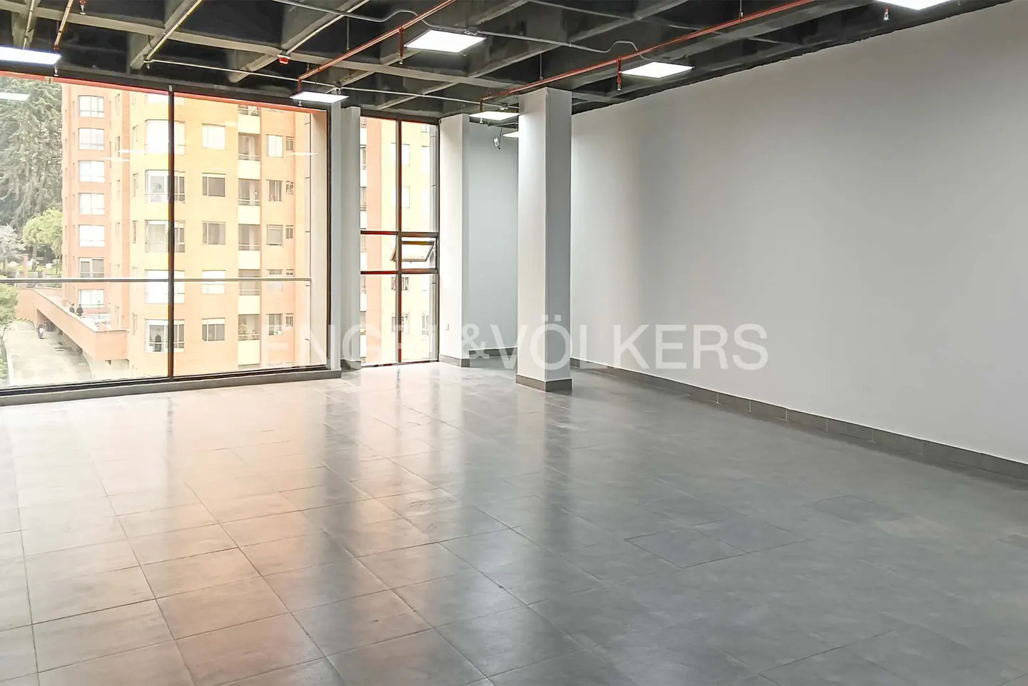 Empty office space with gray tile floor, white walls, and large windows overlooking a building.