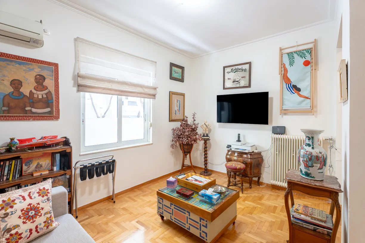 A cozy living room with parquet floors, white walls, and eclectic decor. A colorful coffee table sits in the center, surrounded by art and furniture.