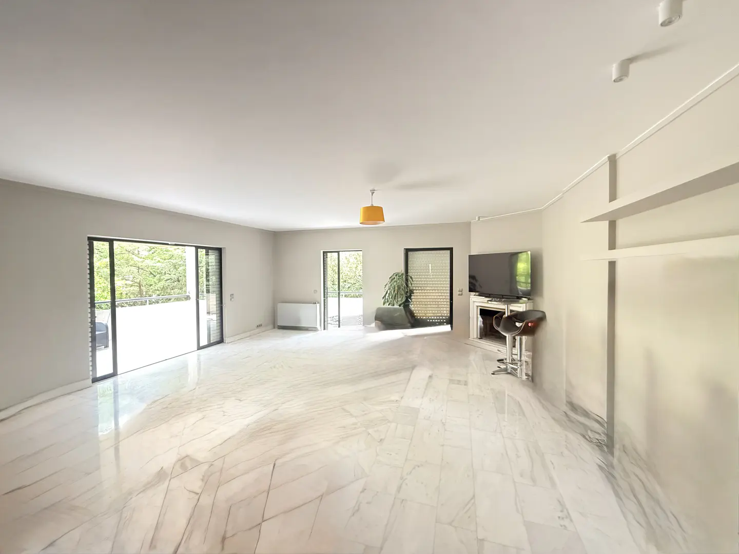 A bright, empty living room with marble floors, sliding glass doors, and a TV over a fireplace.