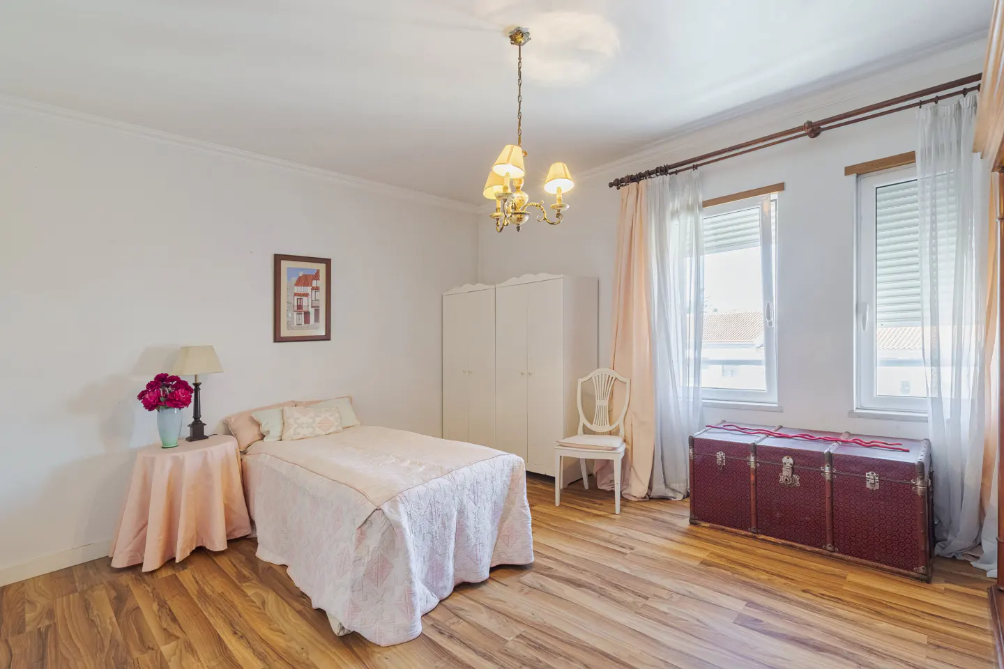 Bedroom with wood floors, white walls, bed with pink cover, white wardrobe, and antique trunk.