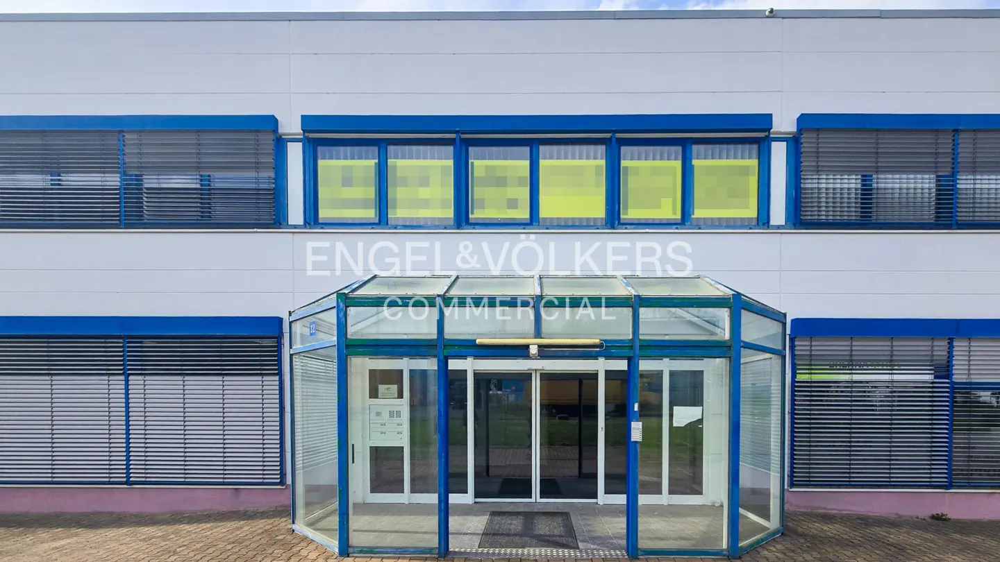 Exterior of a white commercial building with blue window trim and a glass entryway. "Engel & Völkers Commercial" is printed on the building.