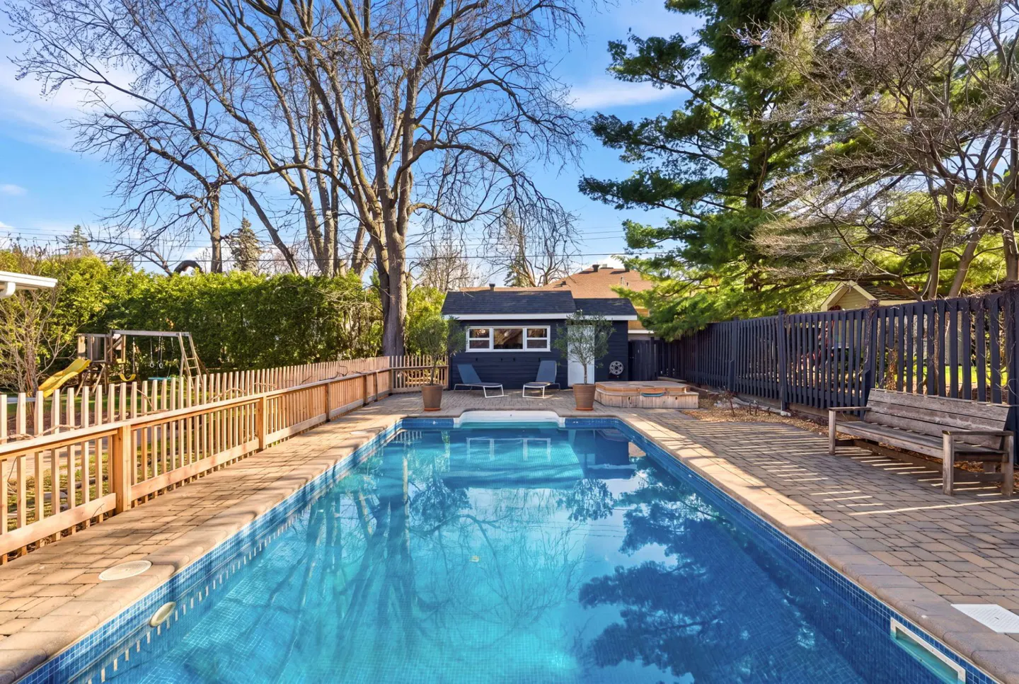 A backyard with a blue tiled pool, a dark blue pool house, a wooden fence, and a bench on a brick patio.