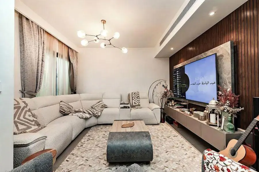 Living room with a white sectional sofa, patterned rug, and a TV mounted on a wood-paneled wall. A guitar leans against a floral chair.