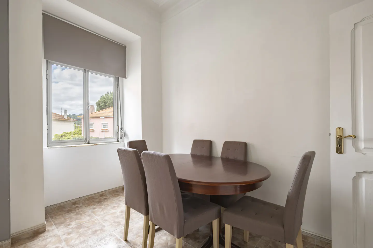 Dining room with a round wooden table and six upholstered chairs. A window with a gray shade looks out onto a residential street.