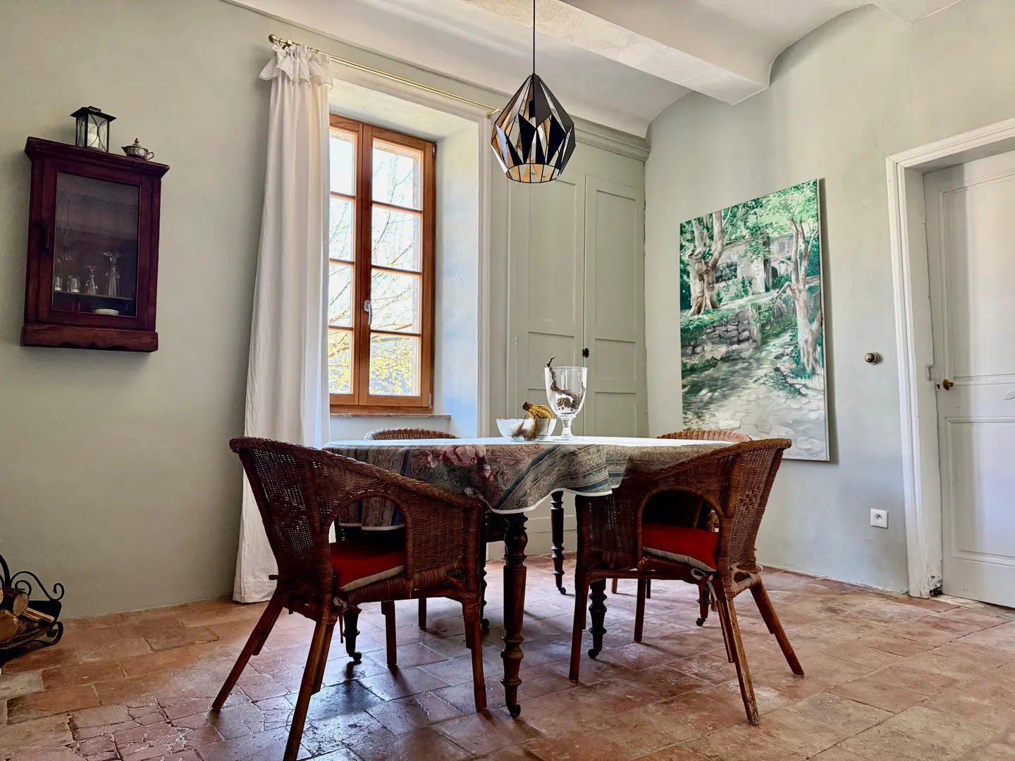 Dining room with brick floors, light green walls, and a wooden table with wicker chairs. A painting hangs on the wall.