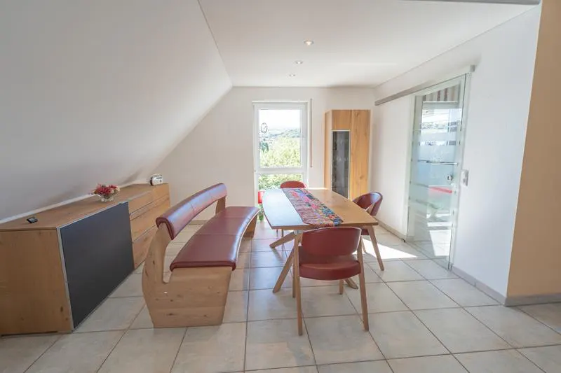 Bright dining room with a wooden table, red chairs, and a bench. A window and glass door let in natural light.