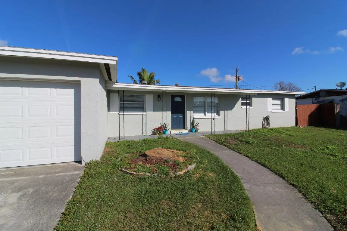 A single-story, light gray house with a dark blue door and white trim under a bright blue sky. A concrete walkway leads to the entrance. A white garage is on the left.