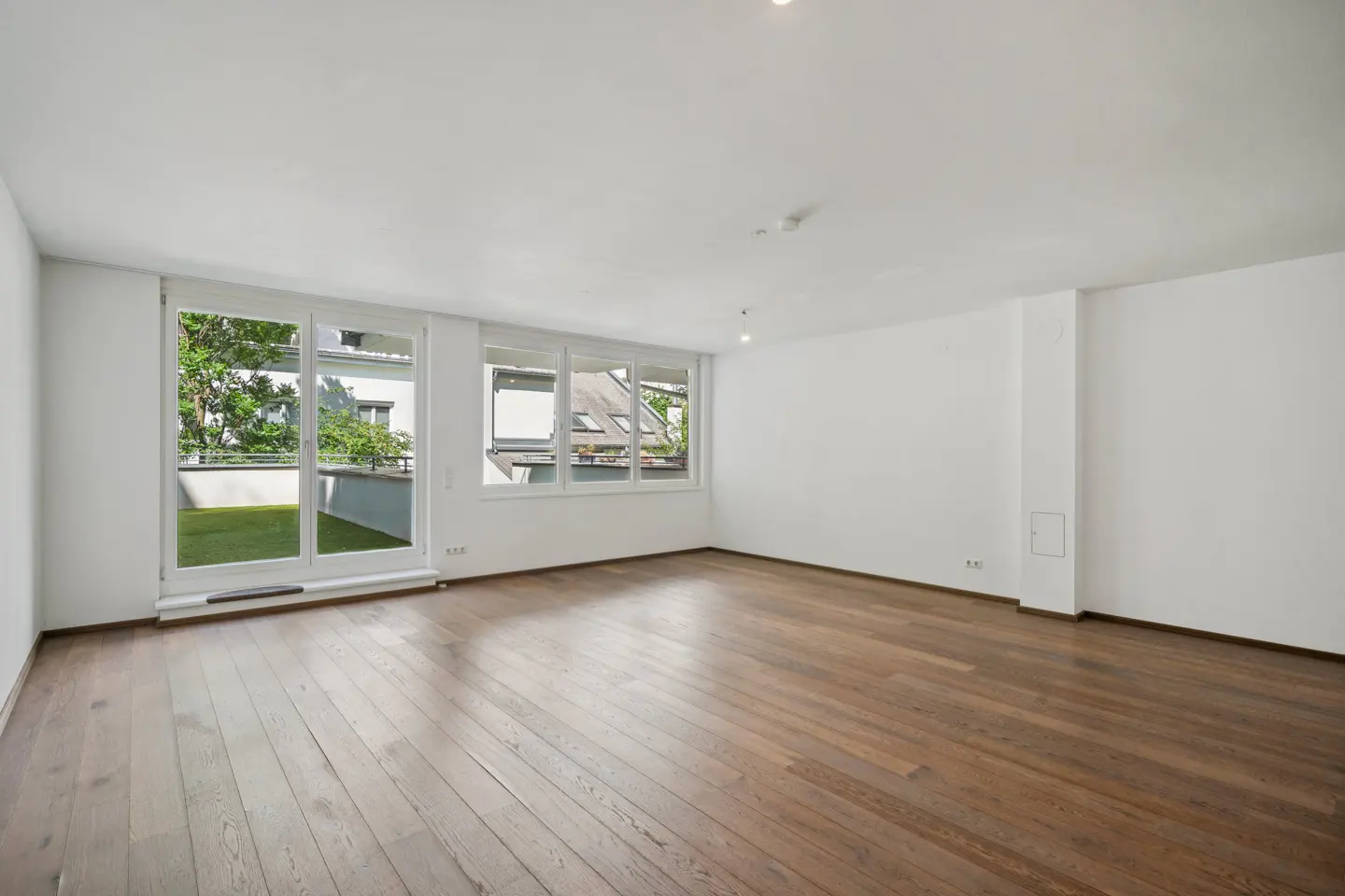 Empty, bright living room with white walls, light wood floors, and large windows leading to a balcony with greenery.