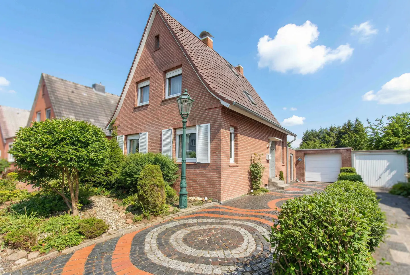 Two-story brick house with a red tile roof, white shutters, and a circular patterned driveway on a sunny day.