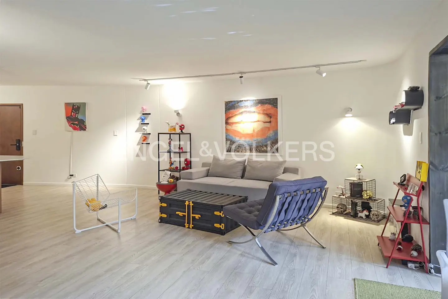 Bright living room with gray sofa, blue chair, and black container table. Artwork and shelving add pops of color. Light wood floors.