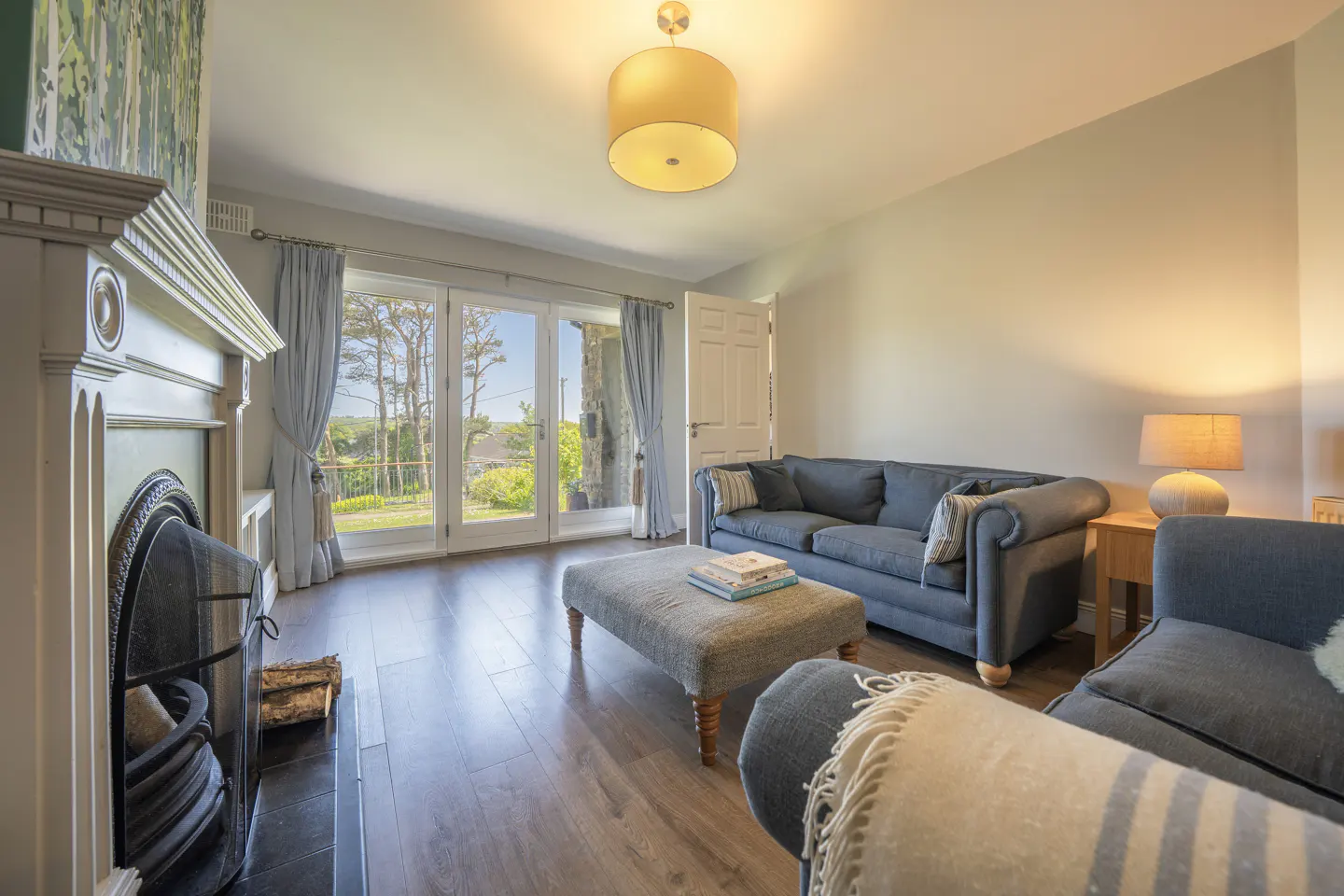 Living room with grey sofas, a matching ottoman, fireplace, and glass doors opening to a sunny outdoor view.