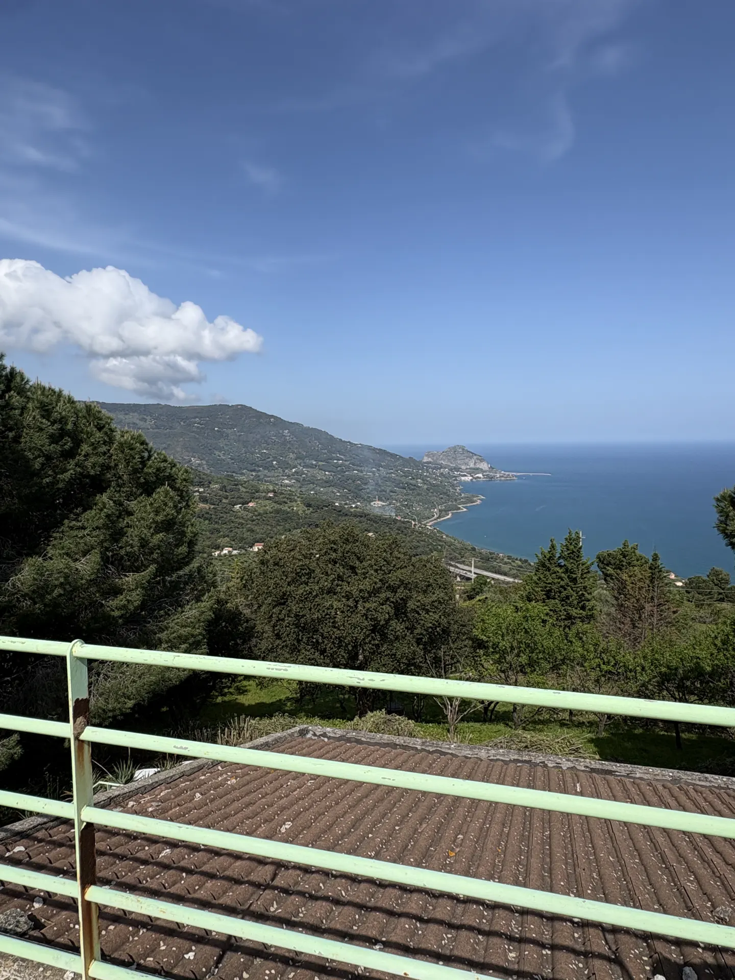 Scenic view from a balcony with green railings overlooking lush hills, a blue sea, and a clear sky with scattered clouds.