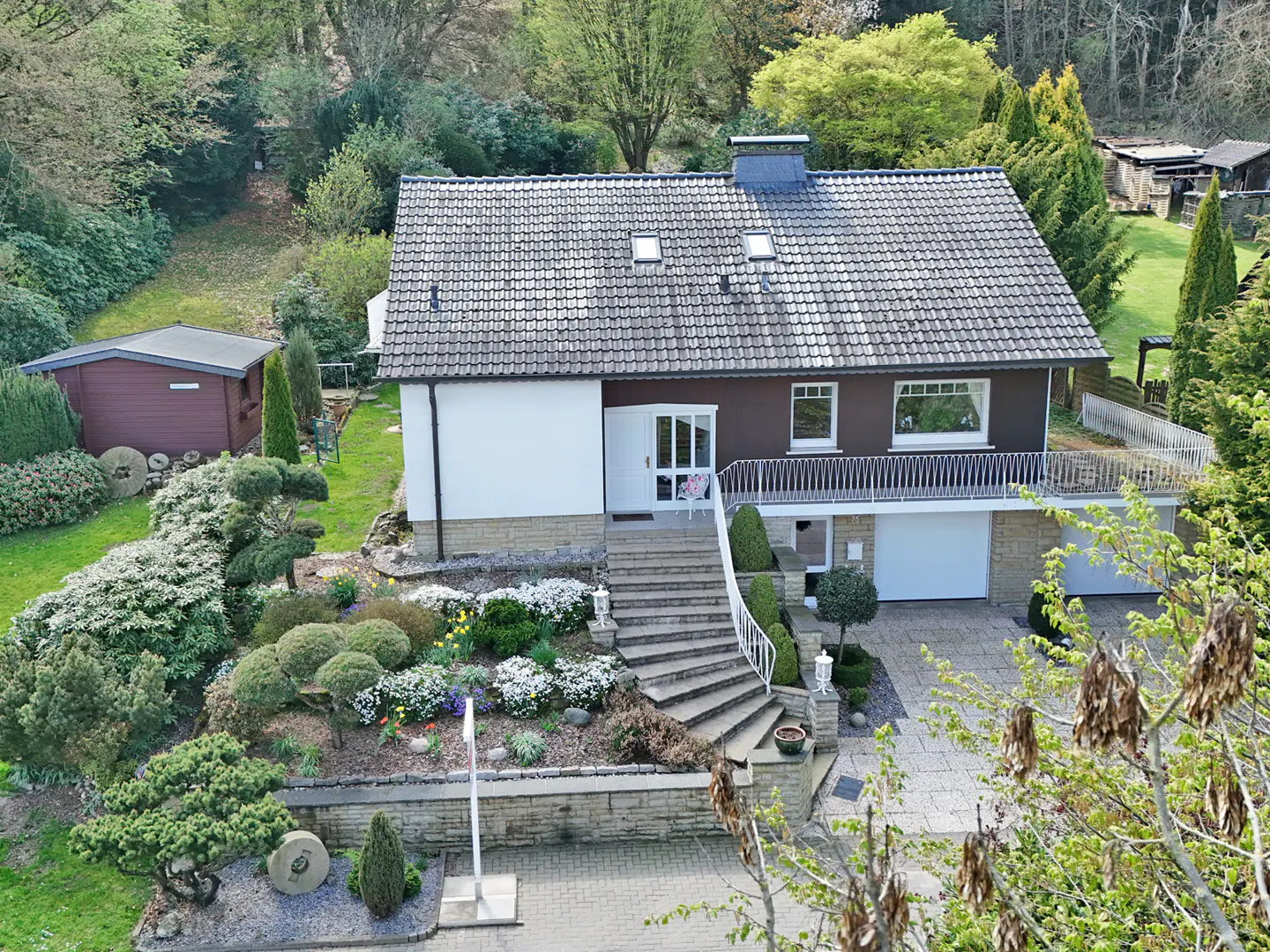 Two-story house with gray roof, brown and white siding, and a stone staircase leading to the front door. Manicured garden and trees surround the property.