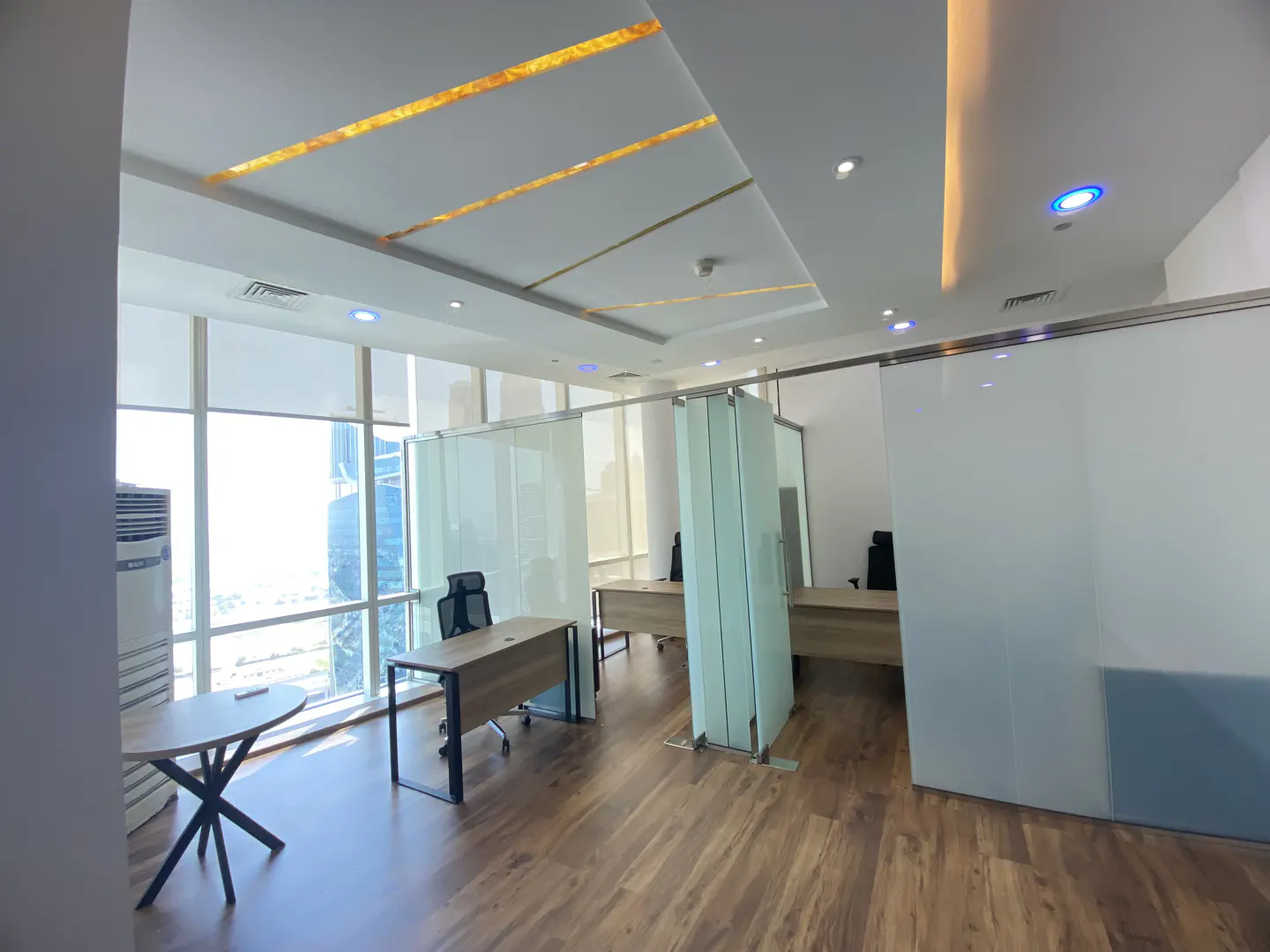 Bright office space with wood floors, glass partitions, desks, and chairs. Large windows offer a city view.