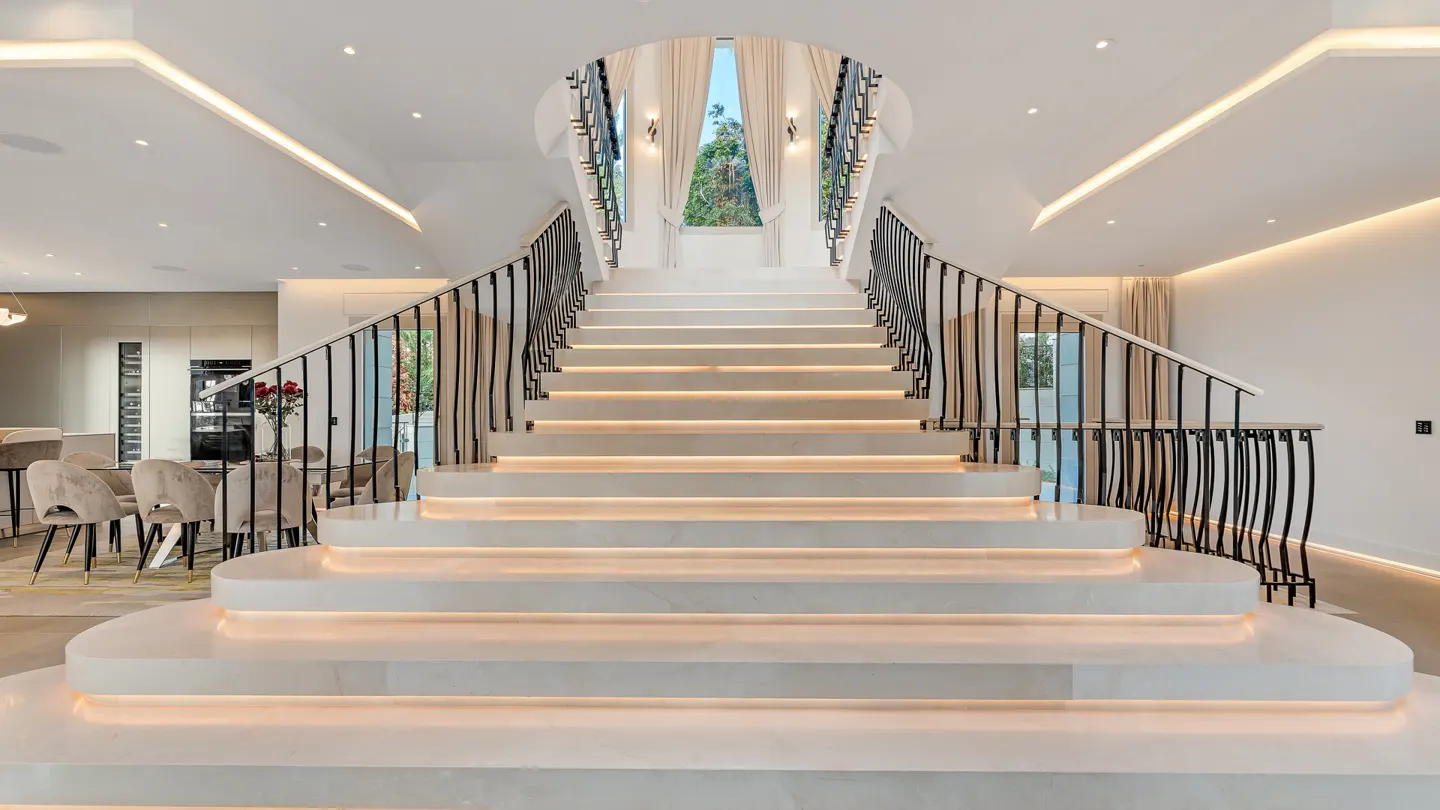 Grand staircase with white marble steps and black wrought iron railings. Steps are lit from underneath. Dining area visible in background.