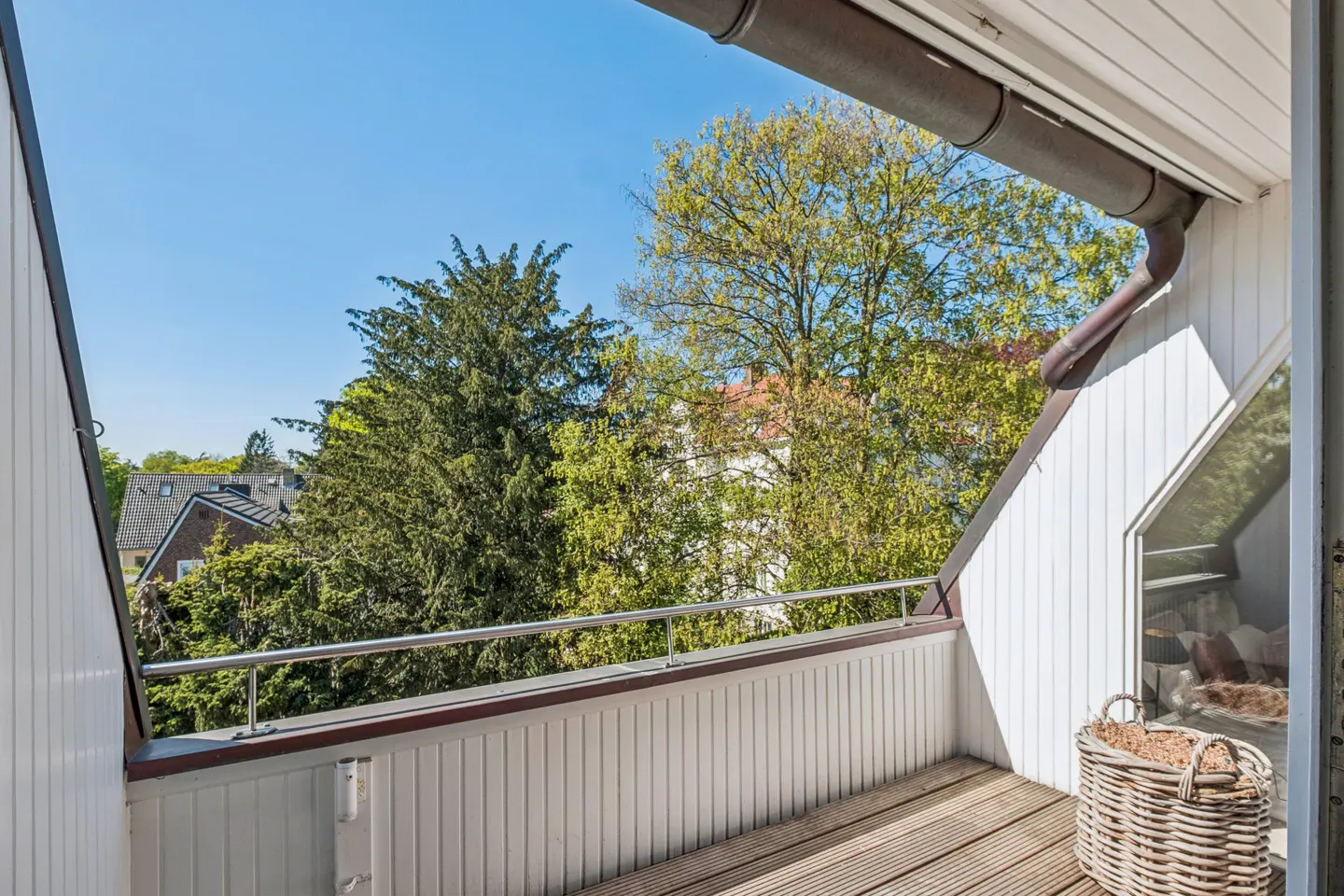 Balcony with wood floor, white walls, and metal railing. Trees and blue sky are visible in the background. A wicker basket sits near the doorway.
