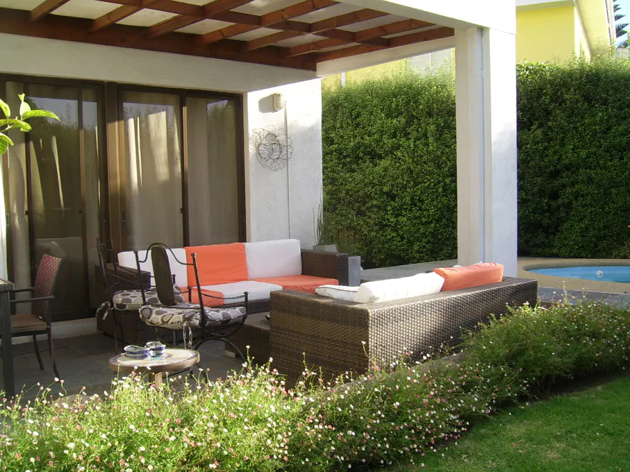 Outdoor patio with wicker furniture, orange and white cushions, and a wooden pergola. Green hedges and a pool are in the background.