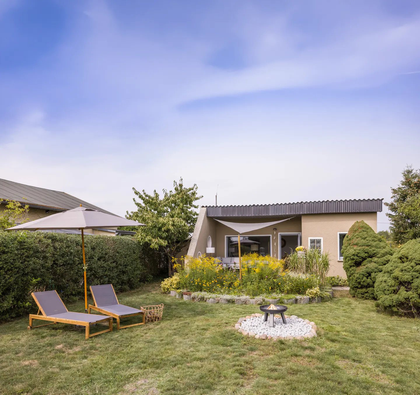 Backyard view with two lounge chairs under an umbrella, a fire pit surrounded by white rocks, and a beige house with a flower garden.