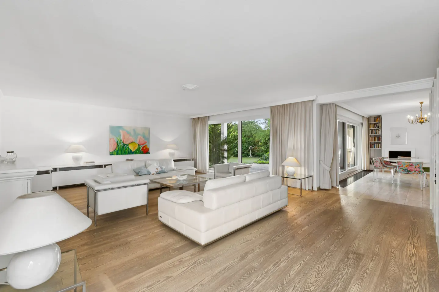 Bright, open living room with white sofas, wood floors, and a view of the garden through large windows. A colorful painting hangs on the wall.