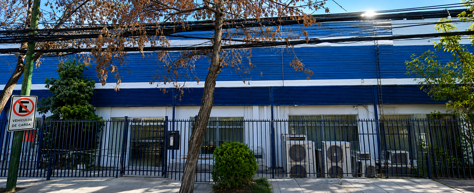 Commercial building with blue metal siding, white ground floor, fenced entrance, and air conditioning units outside.