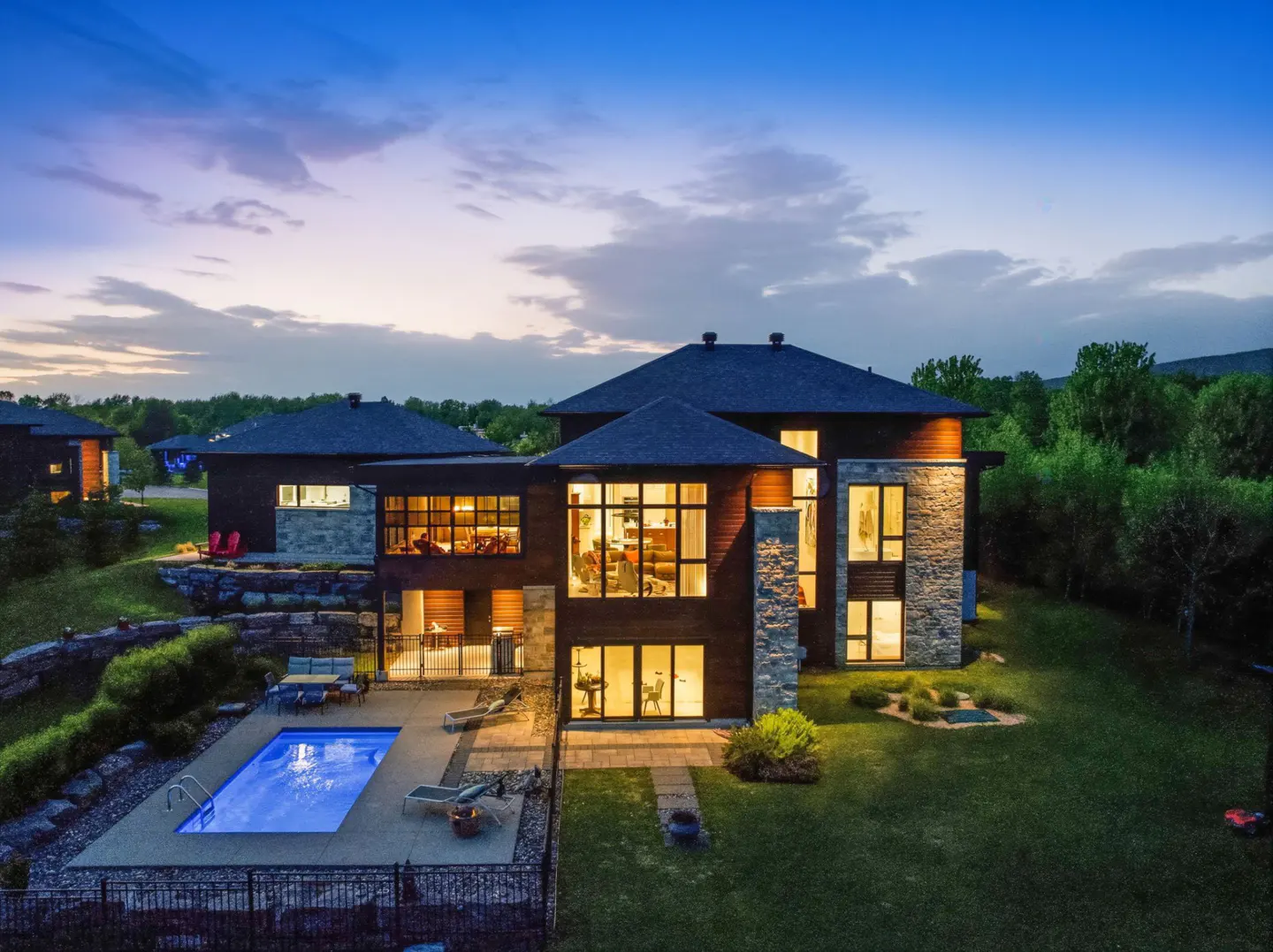 Modern home exterior at dusk with a pool. The house has wood and stone accents, large windows, and a dark roof.