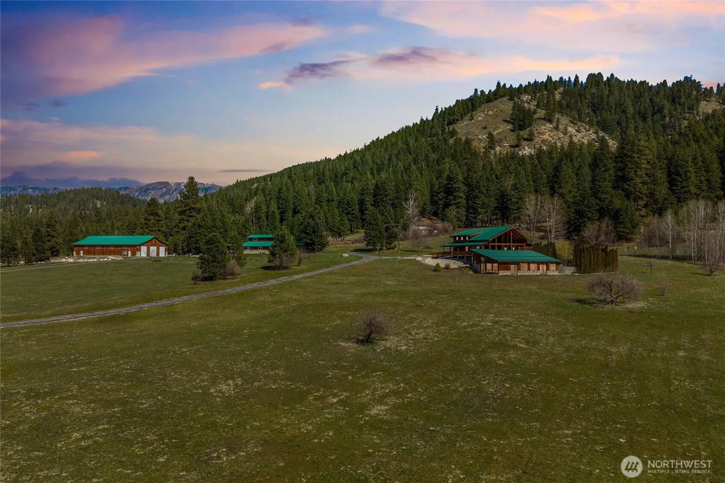 Estate with green roofed buildings on a grassy field, backed by a forested hill and a colorful sky.