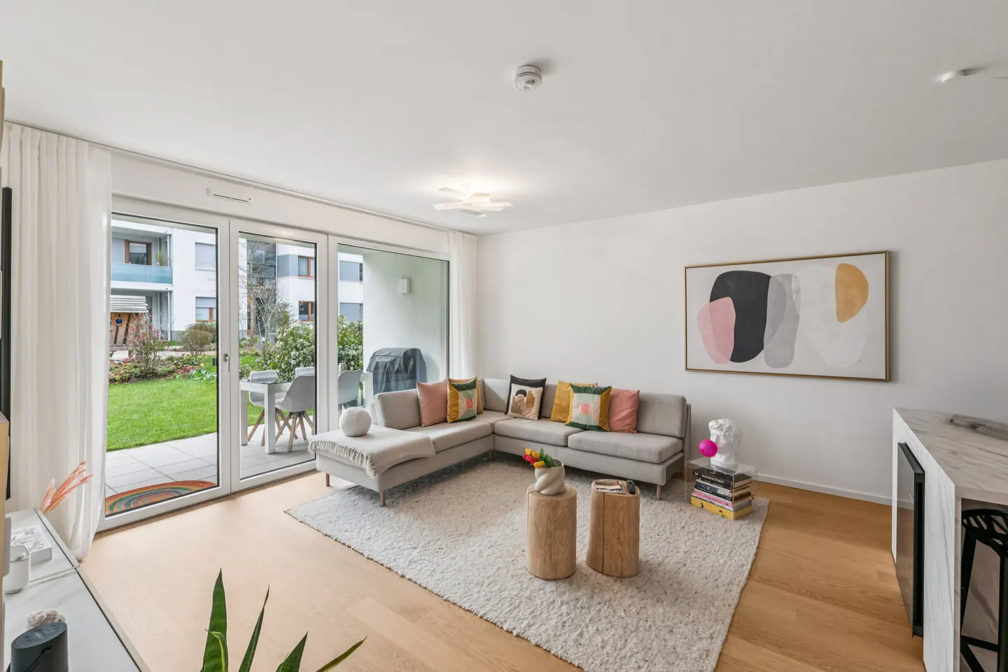 Bright living room with a gray sectional sofa, colorful pillows, and a white shag rug. Sliding glass doors lead to a patio and green lawn. Abstract art hangs on the white wall.