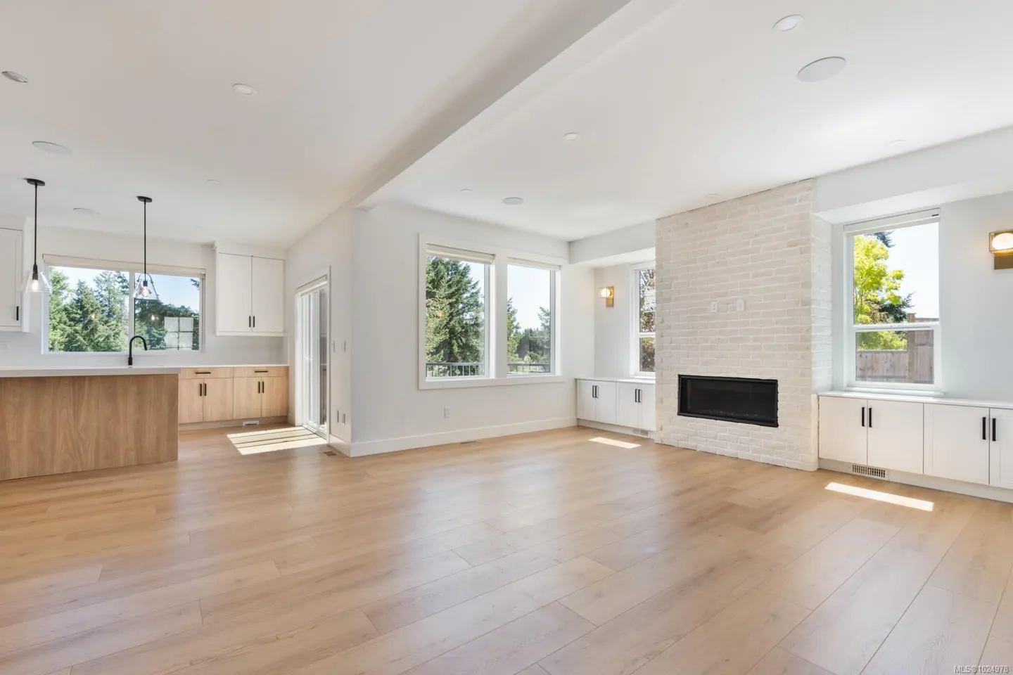 Bright, open-concept living space with light wood floors, white walls, and a brick fireplace. Kitchen island and large windows offer views of greenery.