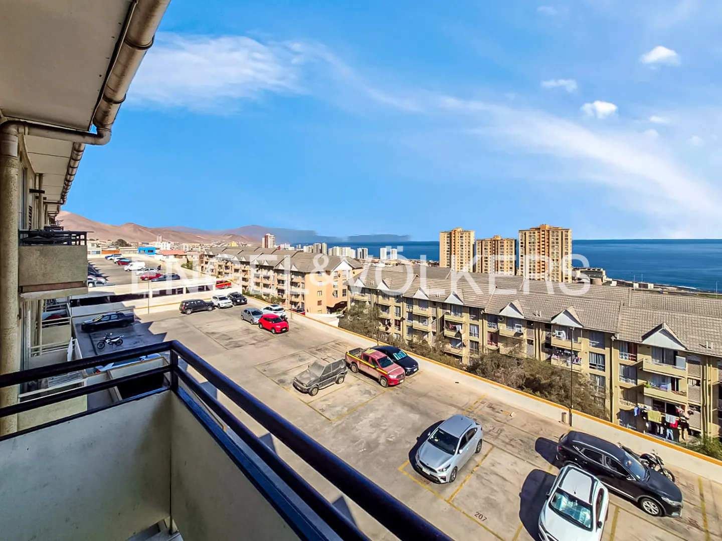 View from a balcony overlooking a parking lot with cars, buildings, and the ocean under a blue sky.
