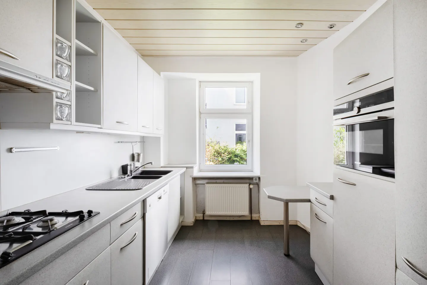 Bright kitchen with white cabinets, dark wood floor, and a window showing greenery outside. Appliances include a stove, sink, and built-in oven.