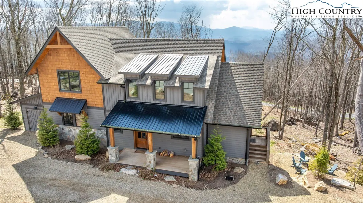 Aerial view of a gray and brown mountain house with a metal roof, surrounded by trees and mountains.