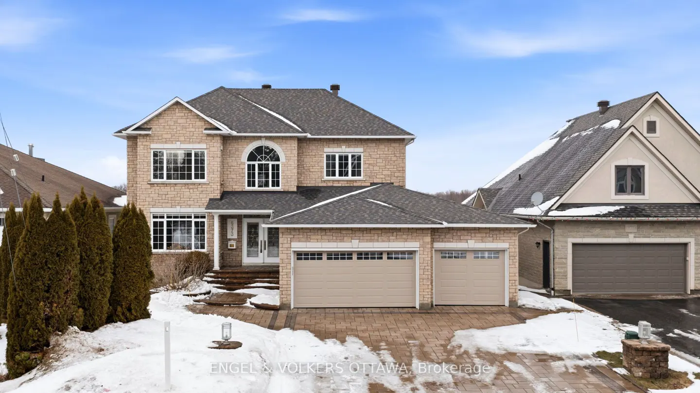 Two-story beige stone house with a gray roof, a two-car garage, and a brick driveway with snow.