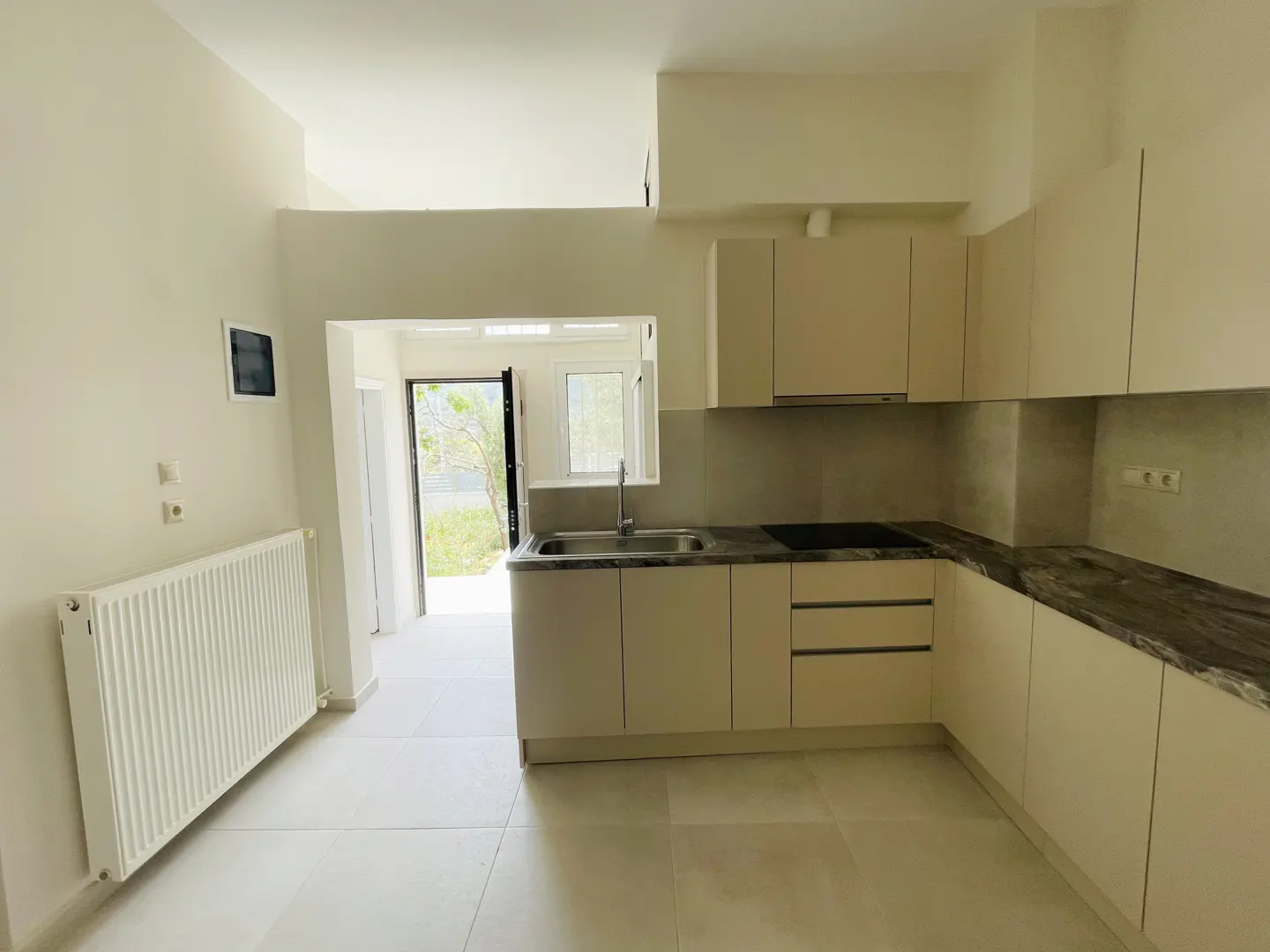 Bright, modern kitchen with cream cabinets, dark countertops, and stainless steel sink. A white radiator is on the left wall. An open door leads outside.