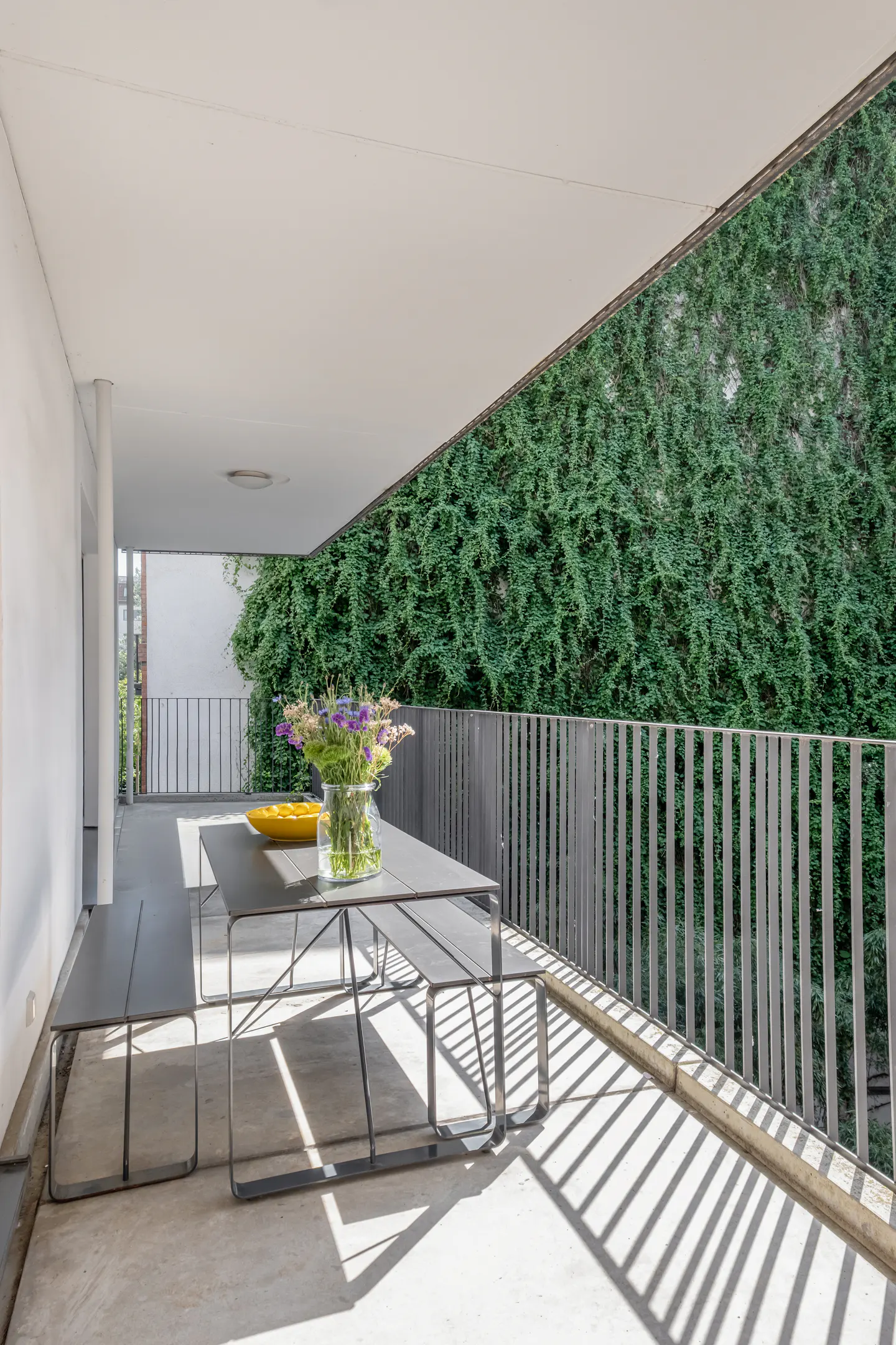 Modern balcony with a dark metal dining set, a vase of purple flowers, and a wall covered in lush green ivy.