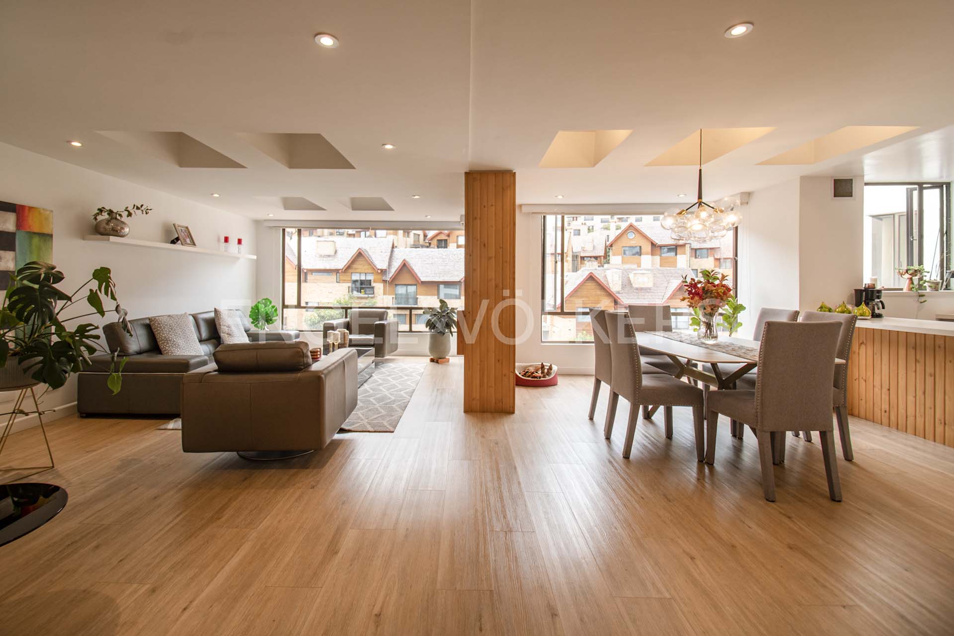 Open-concept living and dining area with wood floors, gray sofas, and a dining table with six chairs. Large windows offer views of houses.