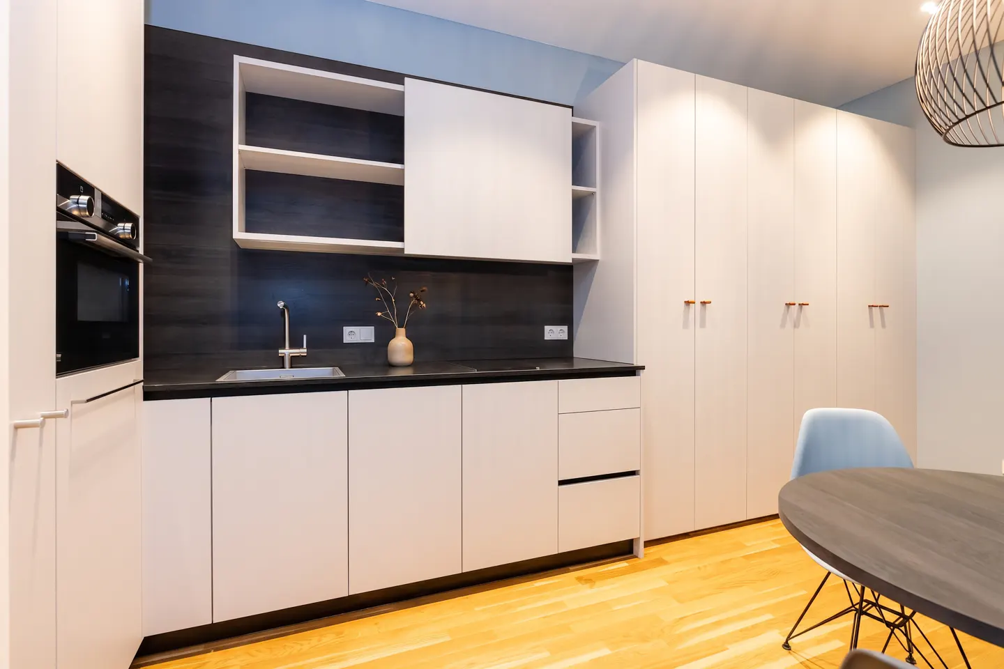 A modern kitchen with white cabinets, black countertops, and a wood floor. A round table with blue chairs is visible.