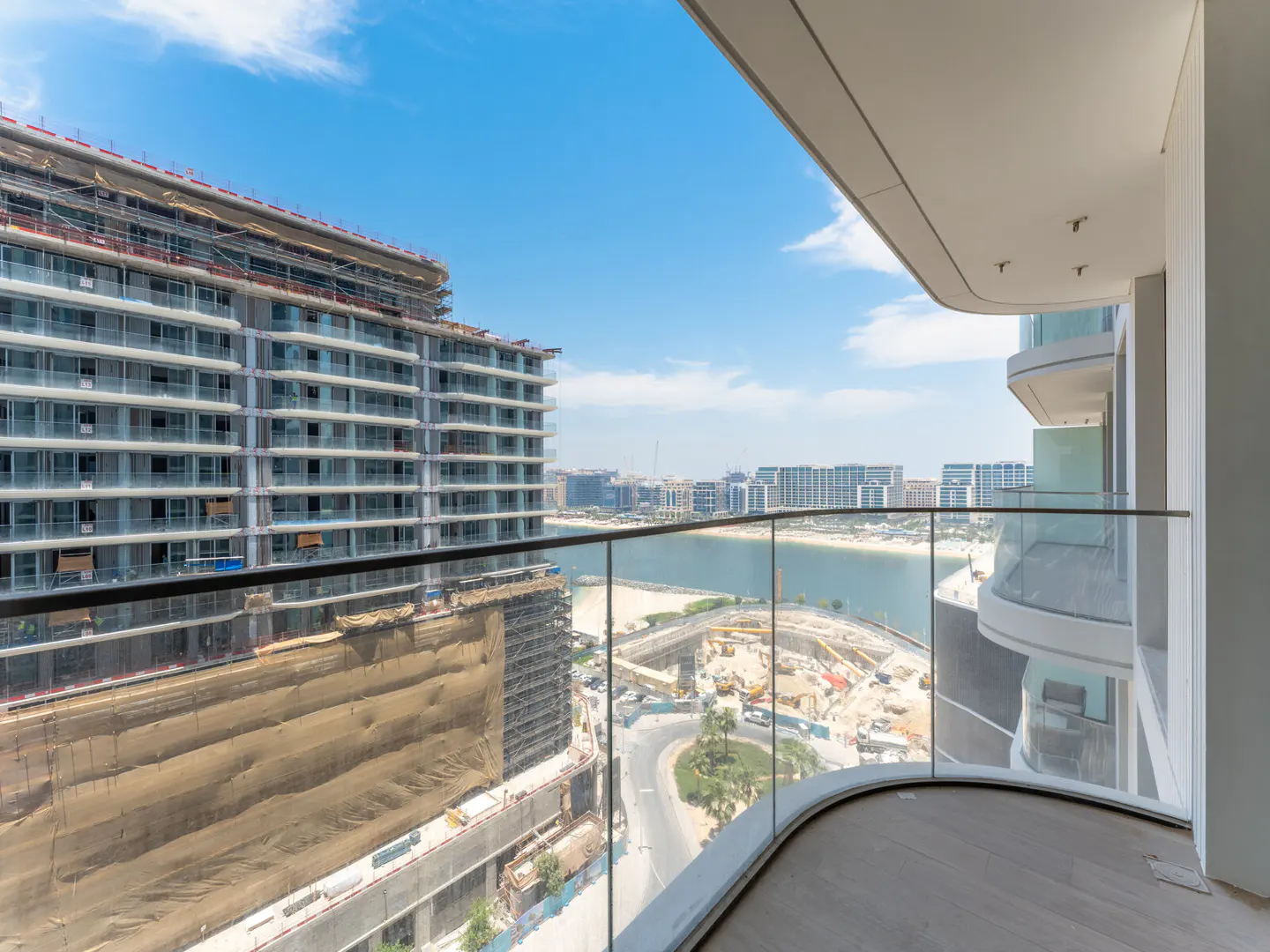 View from a balcony with glass railings overlooking a construction site, a body of water, and city buildings under a blue sky.