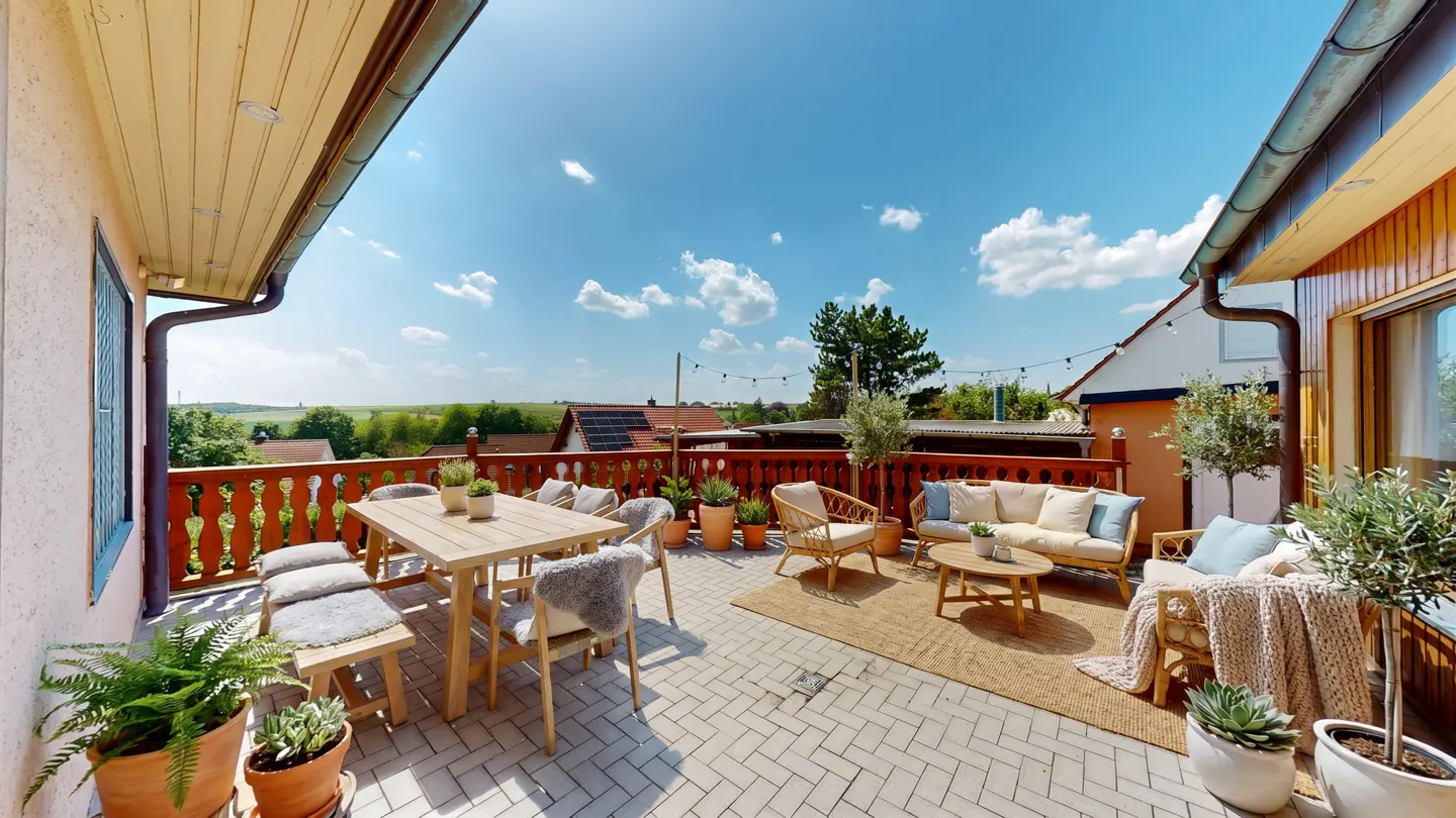 Sunny rooftop terrace with wooden dining set, wicker lounge furniture, potted plants, and a scenic view under a blue sky.